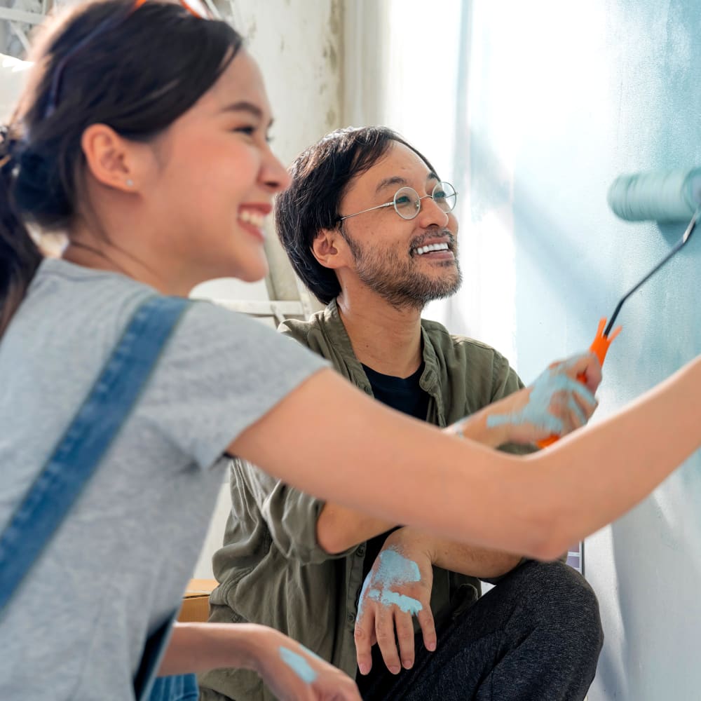 A couple smiling and working together to paint a wall using paint rollers.