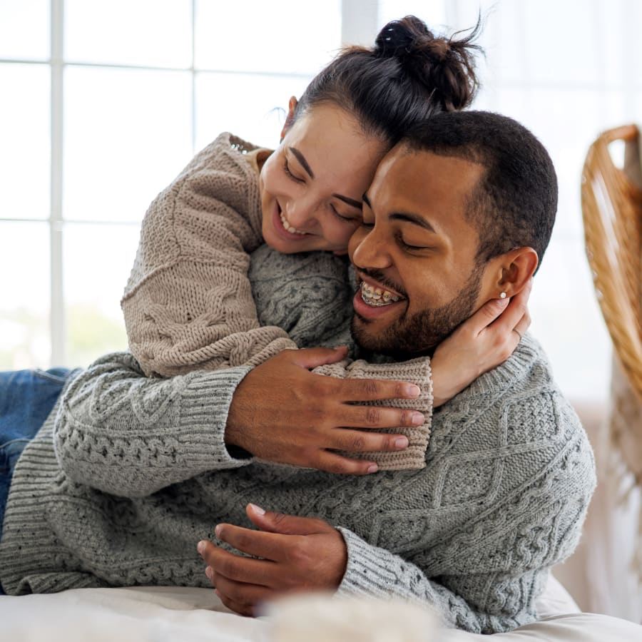 Man with braces smiling as he is being hugged by a lady who is slightly behind him.