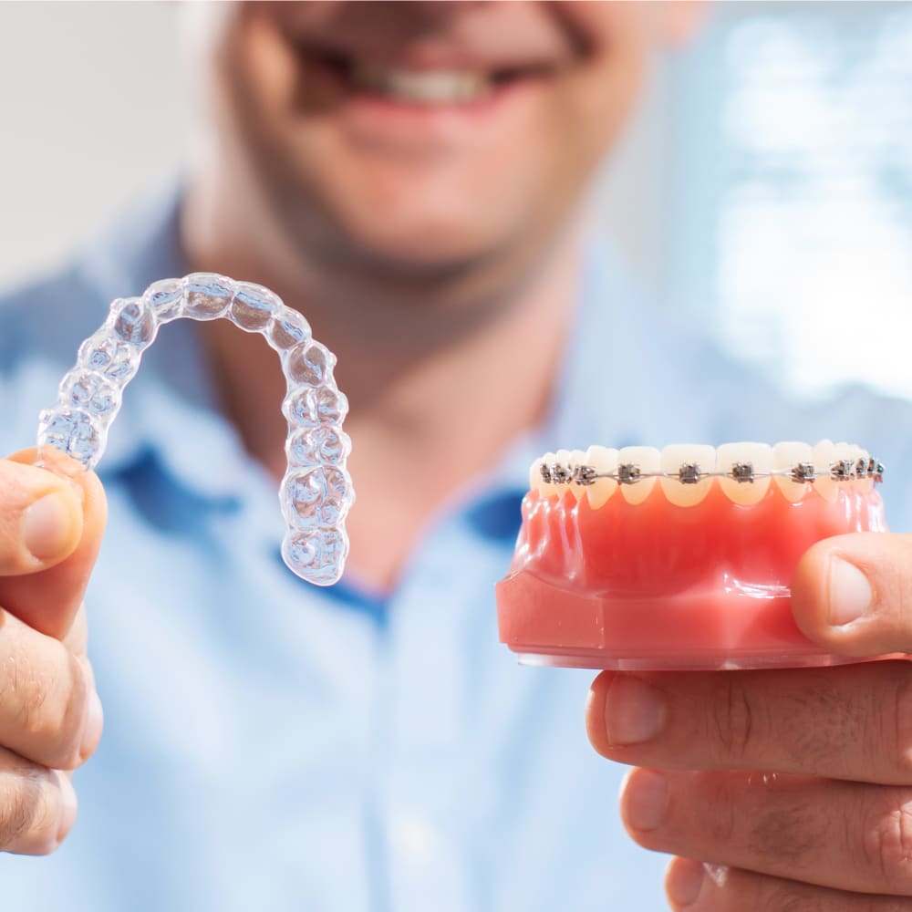 Orthodontist holds up a clear aligner in one hand and model of teeth with braces in the other.
