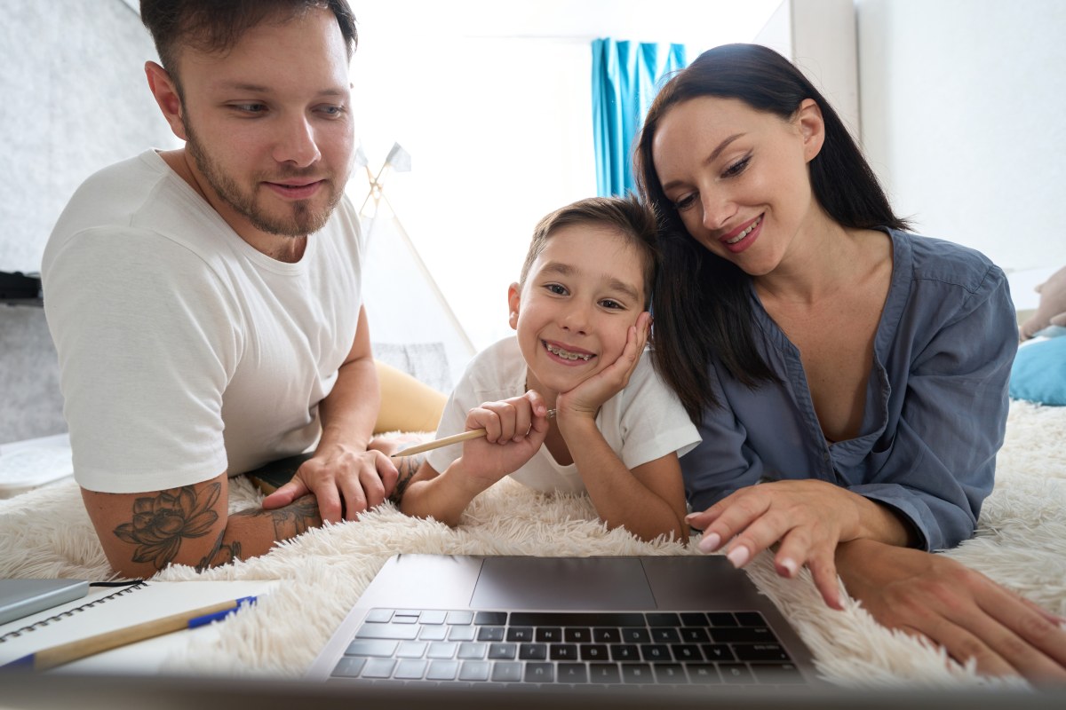 A man, child with braces, and a woman laying on a fluffy carpet together as the boy does homework.
