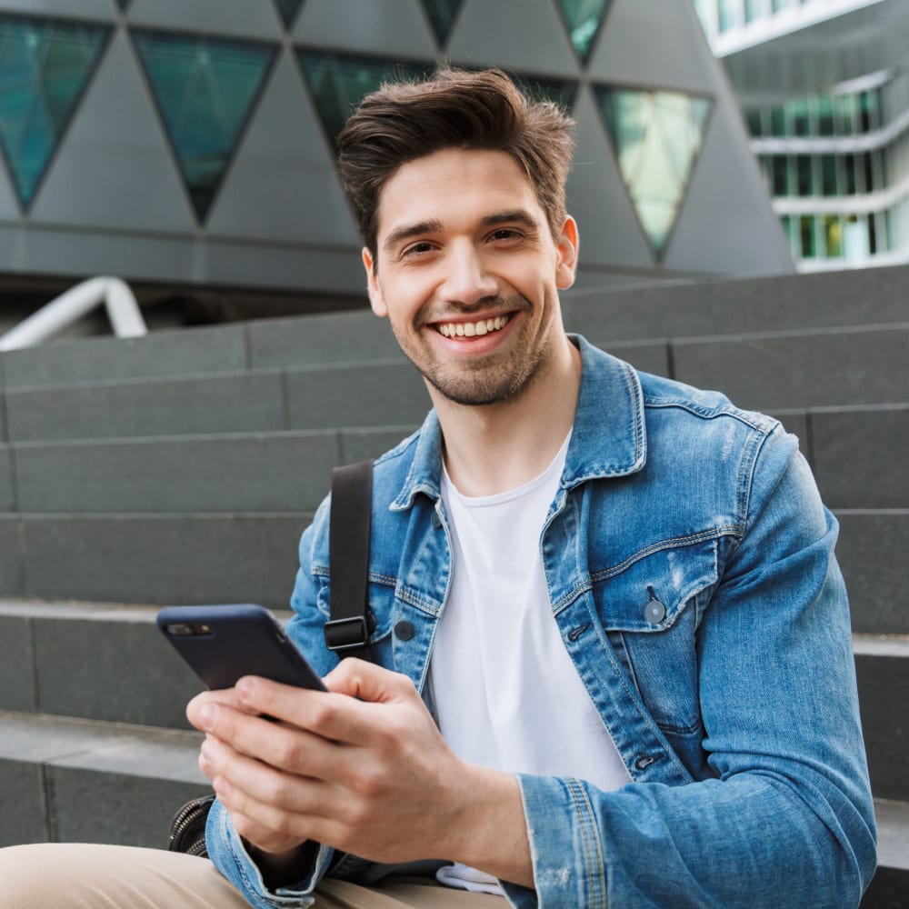 A man in a jean jacket is sitting on stairs and smiling while holding his phone.