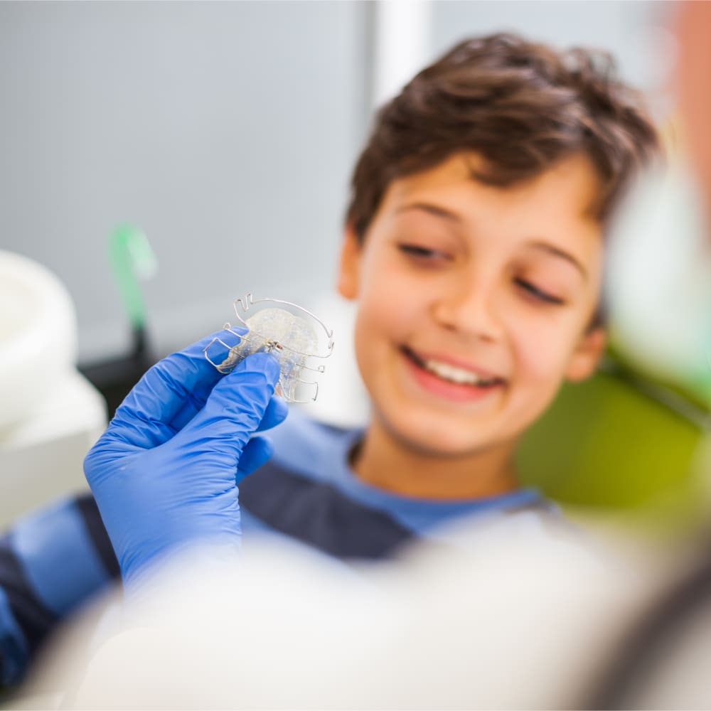 A boy is sitting in a treatment chair and looking at an orthodontic retainer that a team member is holding up.