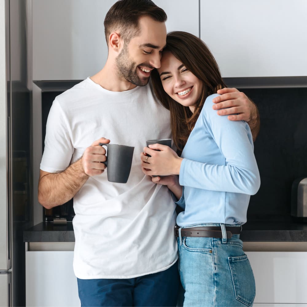 A young couple are laughing and hugging while holding coffee mugs.