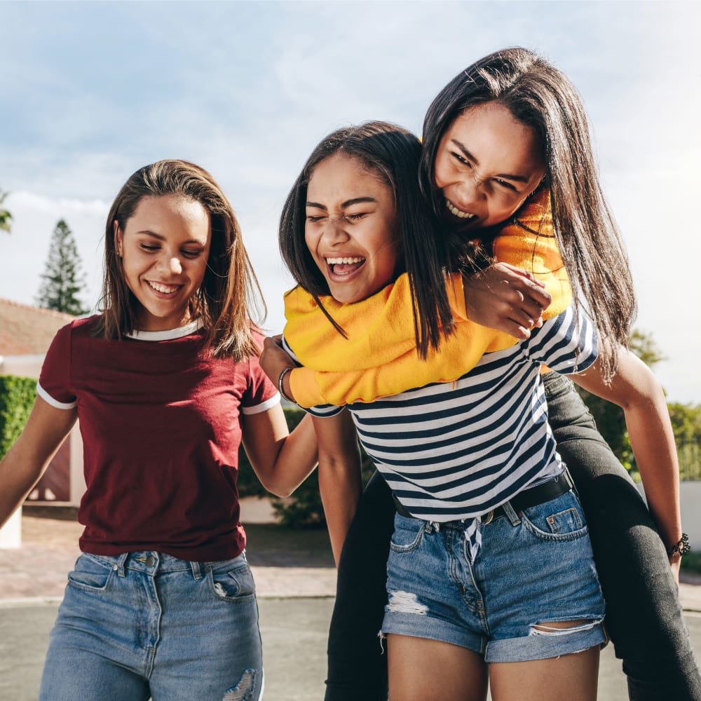 Three teenage girls laughing. One girl is giving another a piggy back ride.