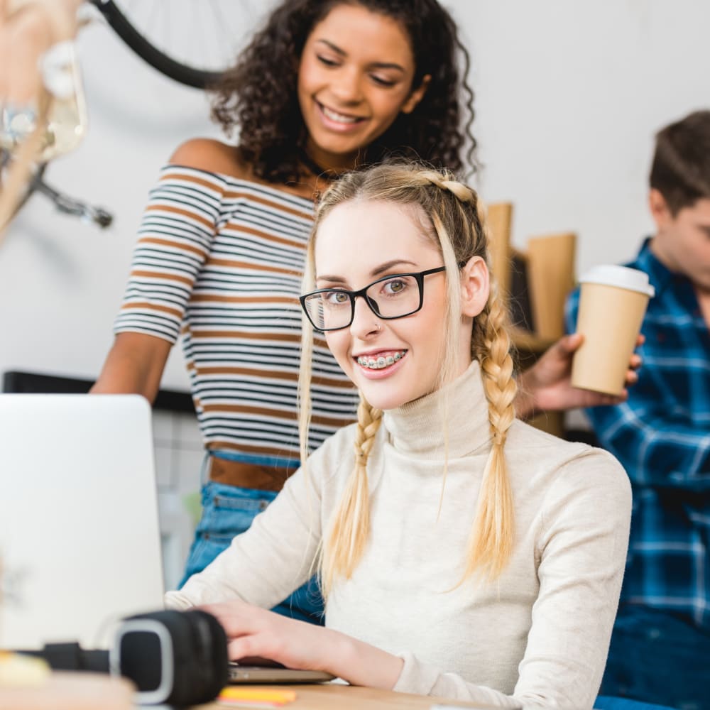 Teen girl with braces and glasses working on a laptop at school with friends around her.